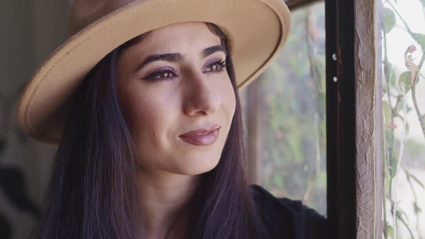A woman wearing a hat sits next to a window with a subtle smile on her face