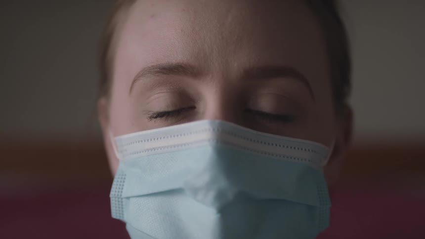 Woman with a ponytail and a light blue mask faces the camera with eye contact