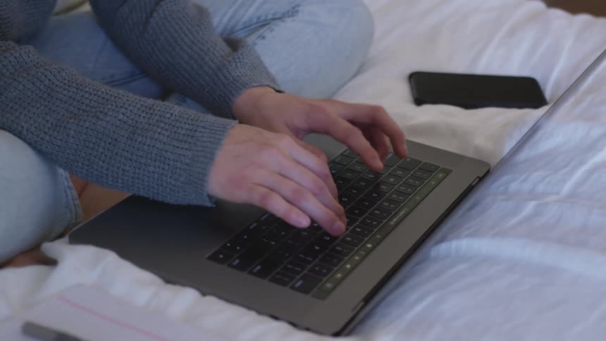 Woman with grey sweater jeans sits on a bed and types on a laptop in the bedroom