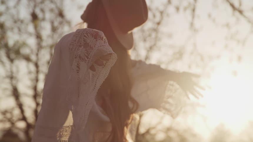 A woman in a sunhat stands under a tree and faces the sky as she stands with her arms held wide