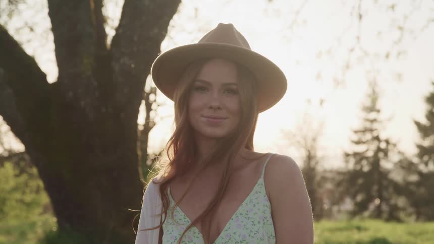 Woman in sleeveless top and hat stands in field near trees with city sprawling in background
