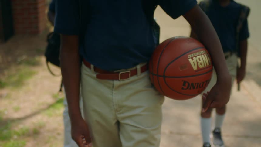 A boy in a school uniform carries a basketball as he leads two other similarly dressed boys down a sidewalk
