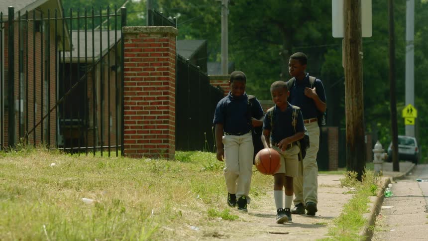 Three boys with backpacks walking on the pavement with one dribbling a basketball