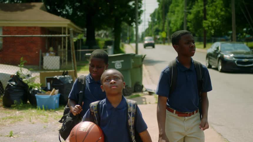 Three boys in school uniforms walk down a neighborhood street as cars drive by