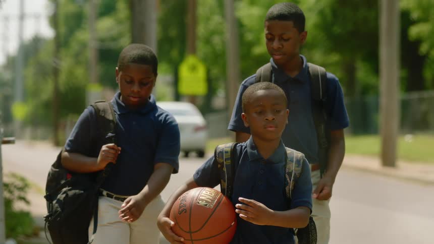 Three boys in school uniforms and wearing backpacks walk down a sidewalk through a neighborhood during the day