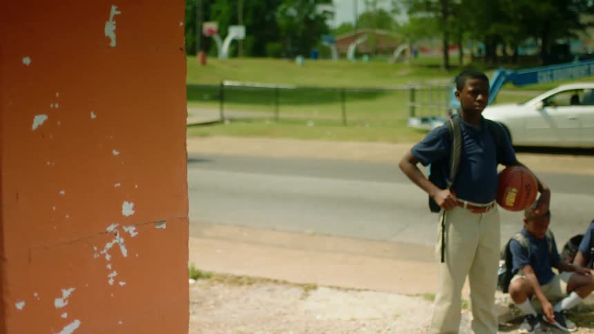 A boy in a school uniform wearing a backpack and carrying a basketball stares into the camera as two other boys sit on the sidewalk behind him