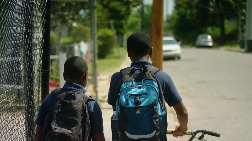 A pair of boys wearing backpacks walk down the sidewalk of a neighborhood as one of them pushes a bicycle along