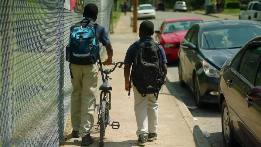 Two boys walk between chain link fence and parked cars one of them pushing a bicycle