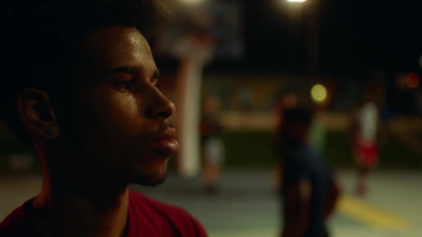 A young black man stands on an outdoors basketball court at night while lost in thought