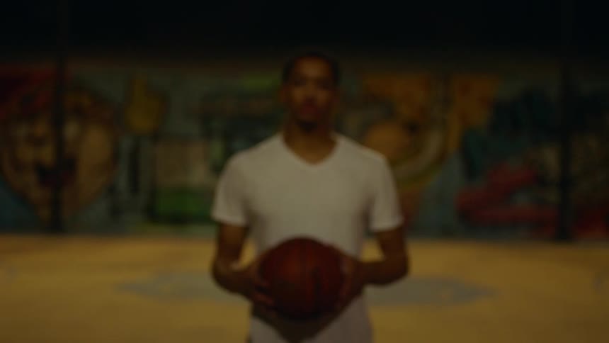 Black man stands and holds a basketball in front of graffiti walls in a basketball court at night