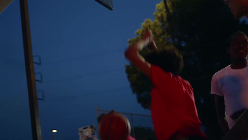 A group of teens plays basketball on an outdoor court as one boy jumps up and puts the ball in the basket as he holds on to the hoop