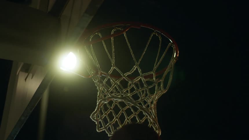 A basketball being shot into a basketball hoop with a streetlight and the sky in the background