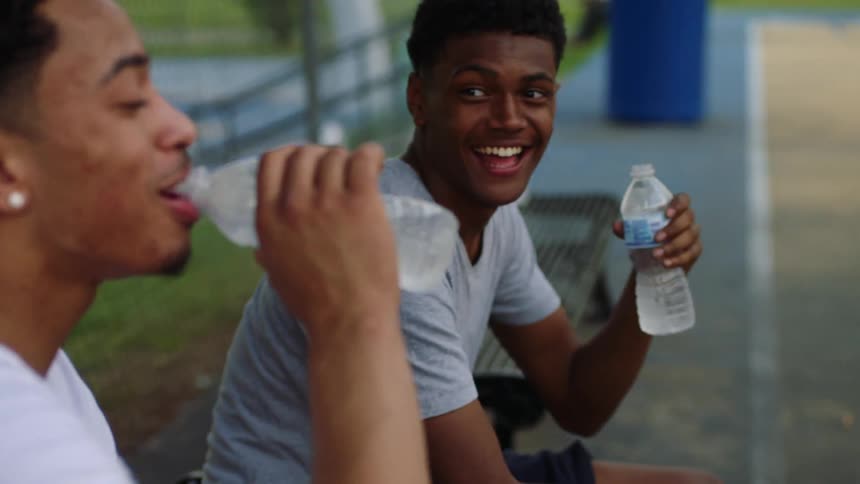 Couple of friends sitting on a bench drink water from plastic bottles while talking and laughing