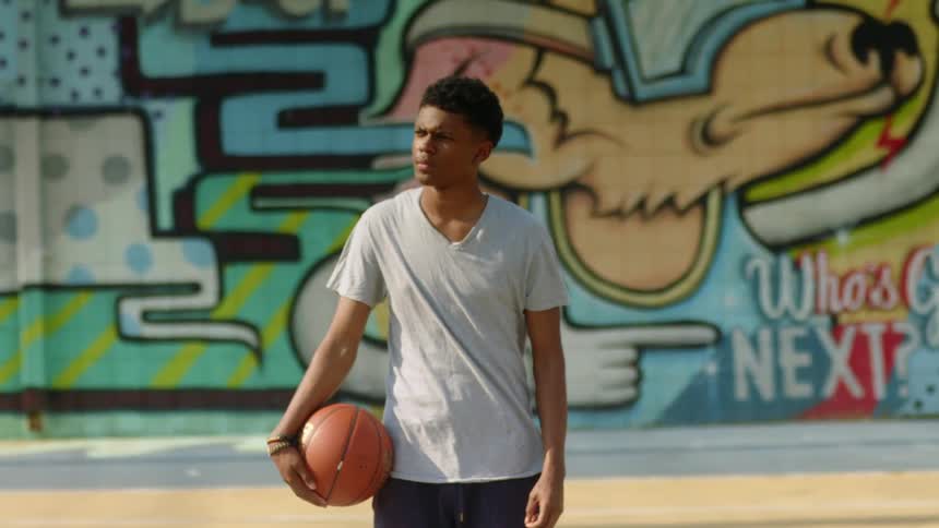 Teenage black boy holds a basketball and stands in front of a graffiti wall in the court