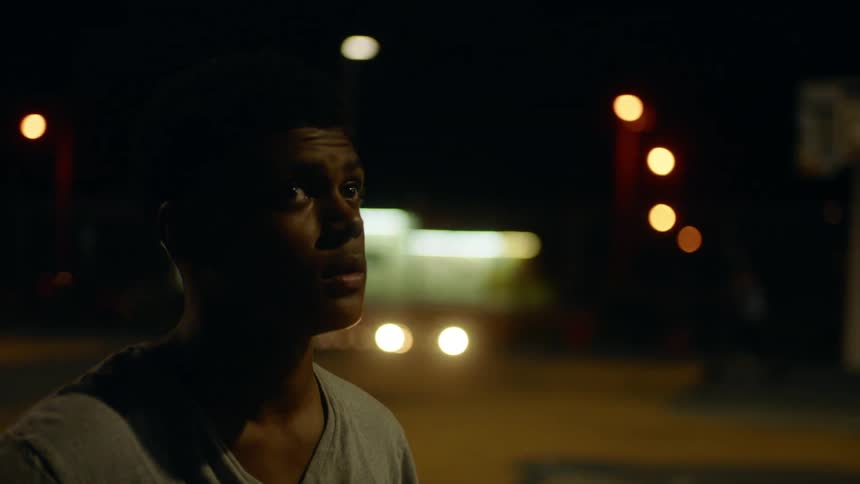 A young man holding a basketball gazes at his surroundings while standing on an outdoor basketball court at night