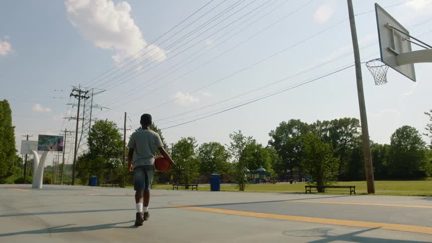 Black boy with shorts white socks boots and a grey t shirt holds a basketball and walks in the basketball court