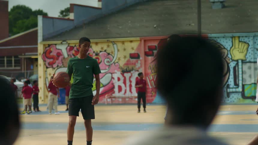 Teenage boys tossing a basketball and a group of young kids stands in the background and others watch the game