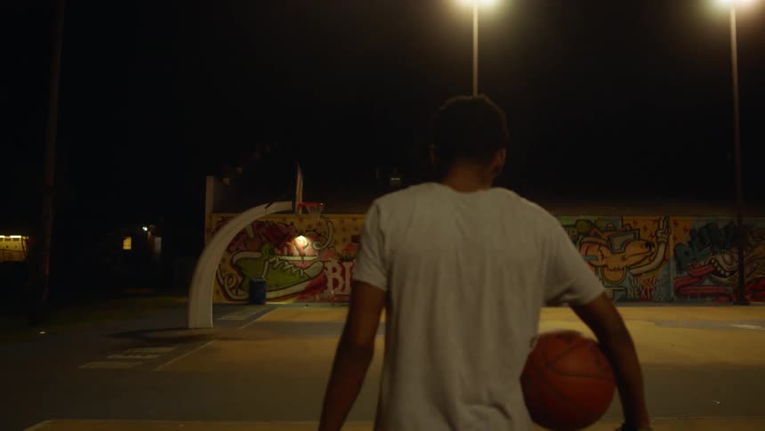 A teenage boy walking across an outdoor basketball court carrying a basketball