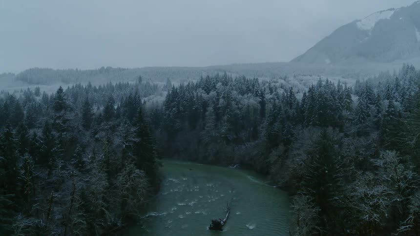 A river valley is surrounded by thick woods covered with frost as snow flurries fall from a gray sky