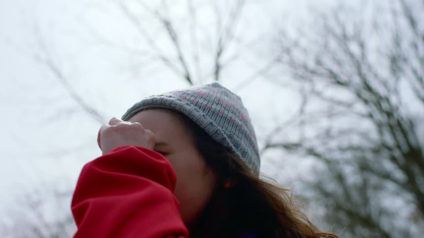 A girl in a jacket and knit cap standing outdoors makes a telescope with her hand and looks around the area