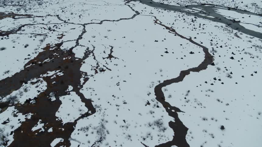 Winding icy creeks flow through a snow field into a cold mountain lake in the distance