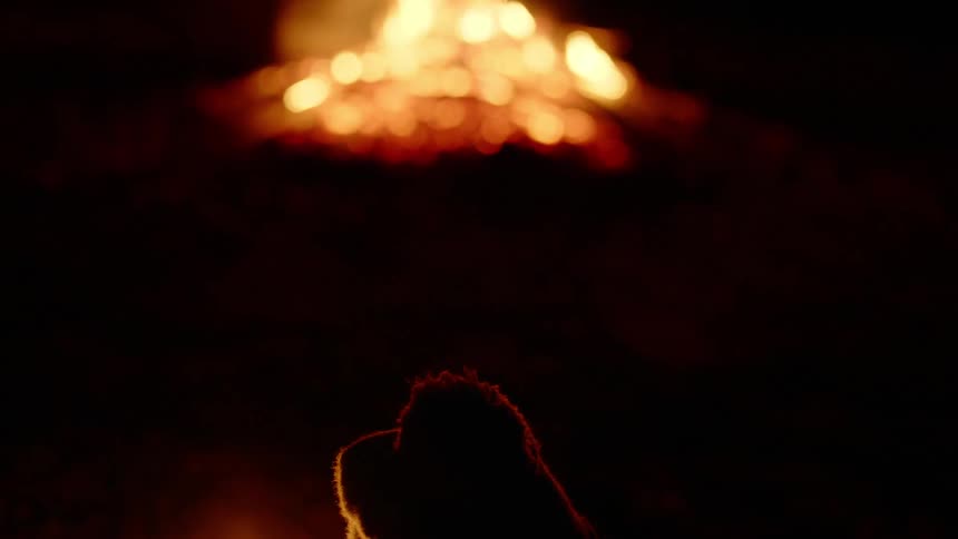 Person sitting in front of the flames of a campfire at night