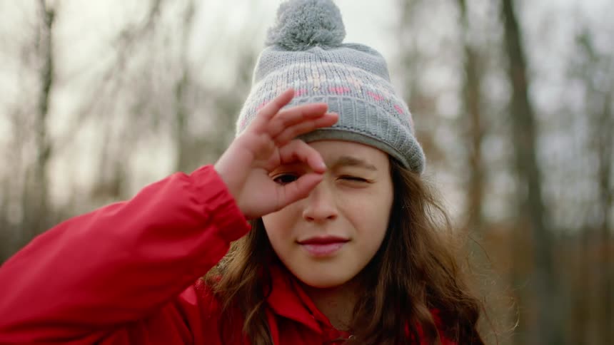A young girl holds her fingers over her right eye like binoculars while posing near a forest