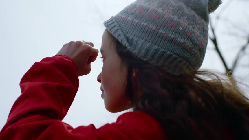 A girl in a hat and red jacket makes a telescope with her hand and looks through her fingers