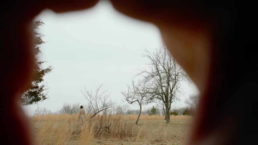 Person focuses through hands at woman standing in field of dried grass