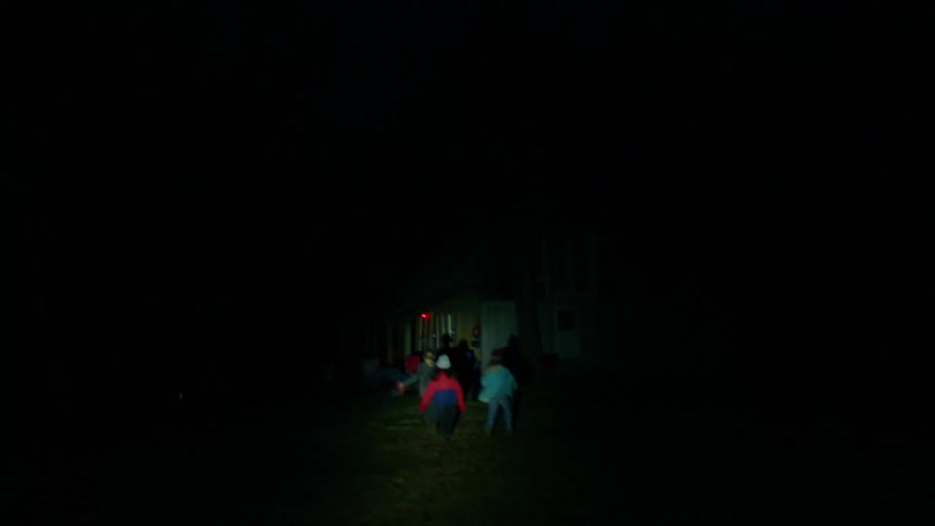 Three girls illuminated by a flashlight run playfully through a yard towards a house at night