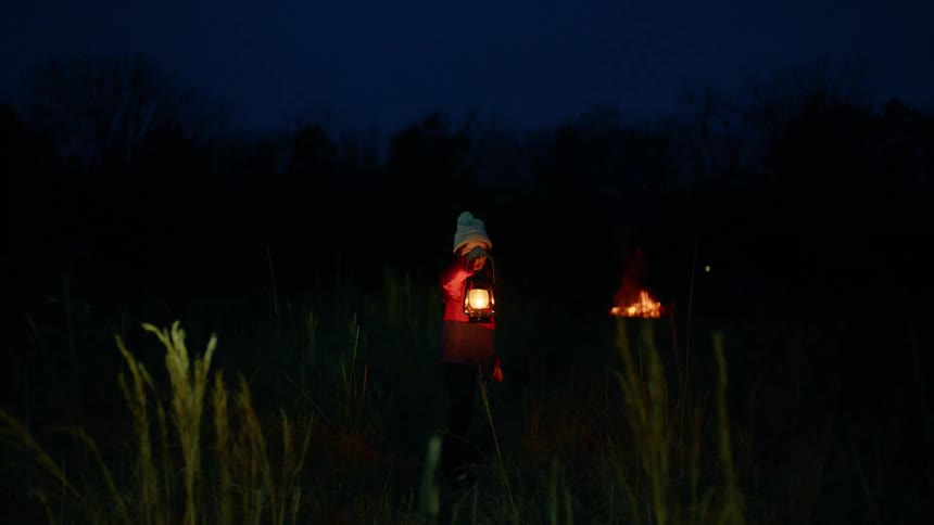 A young white girl is holding a lantern and walking away from a bonfire in an outdoor environment at night