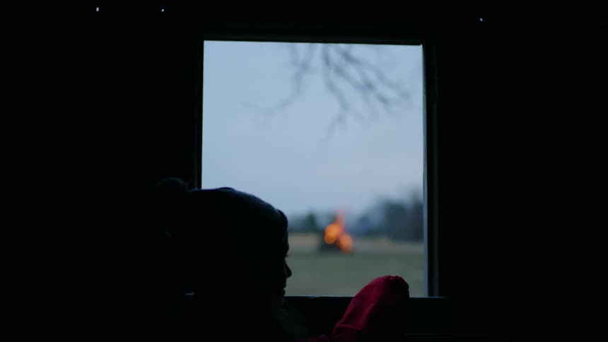 A girl standing in a dark room looks out the window at a large bonfire in field outside