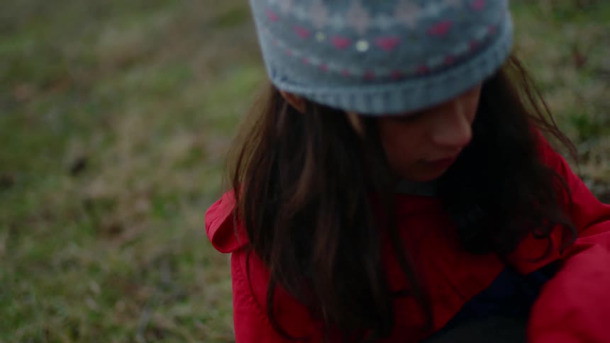 A girl with a hat and jacket sitting on grass holding and stroking a cat