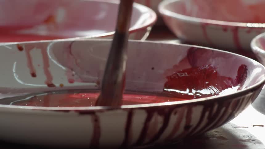 A paintbrush mixes red paint in a white ceramic bowl