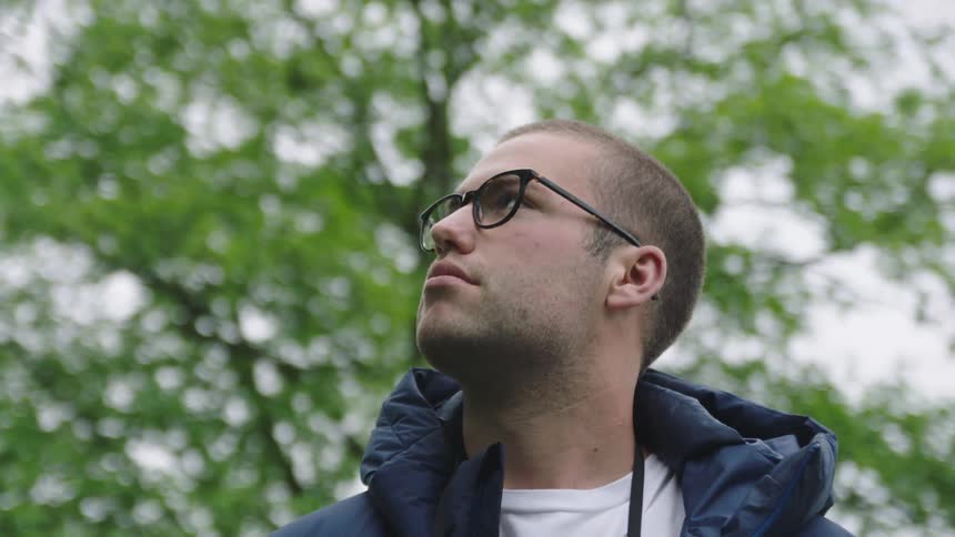 A man is standing outdoors while wearing a coat and glasses