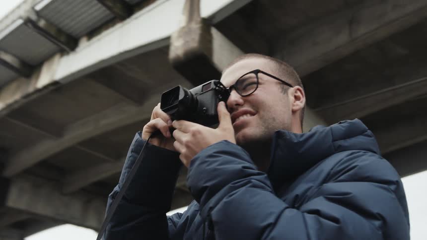 A man holds a camera to his face standing under a bridge