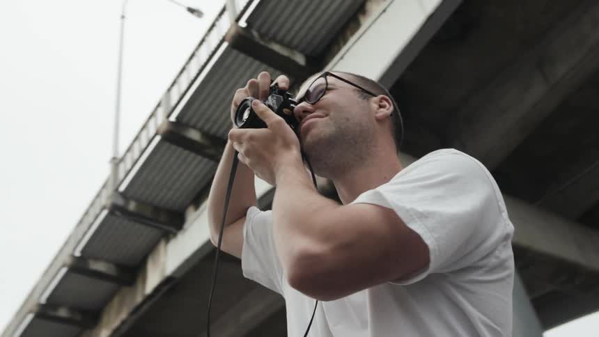 A young man standing beneath an overpass raises a small camera to his eye and aims it to take a photograph