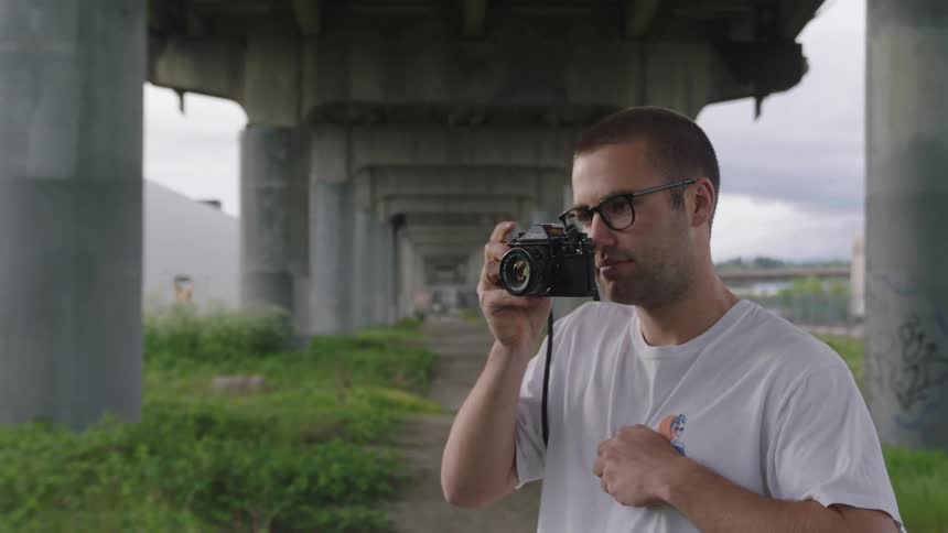 A man stands under a bridge near an ocean and holds up a camera to his face