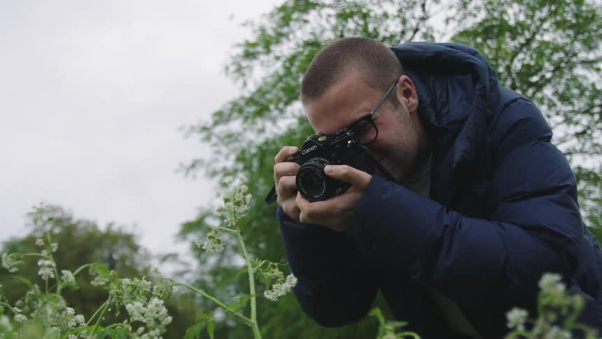 A photographer wearing a blue puffer coat takes close up photos of small white flowers