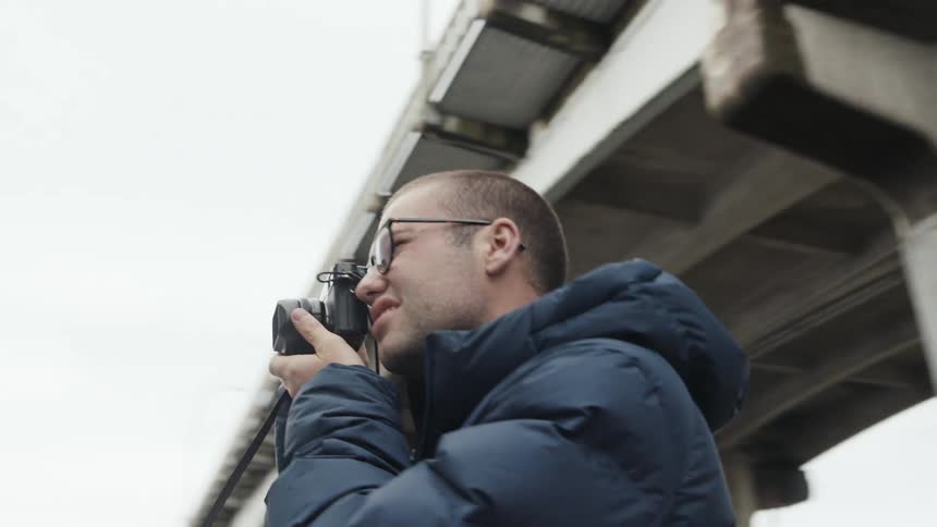 A man holds a camera to his face standing outside underneath a bridge