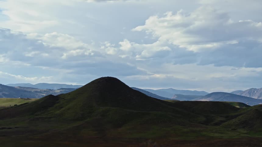 Low green mountain stands against blue and grey mountains on the horizon under a cloudy sky