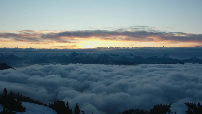 Sun on the horizon turns sky orange as snow and conifer covered mountain peaks peek over the cloud layer