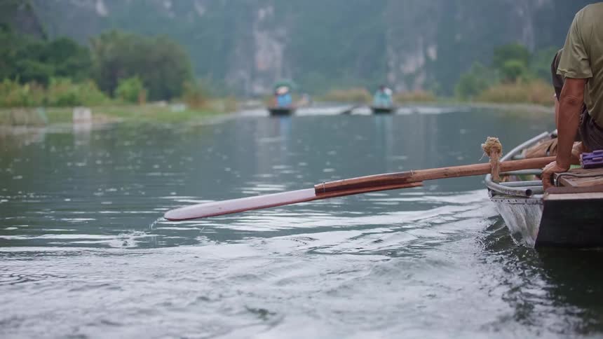 A man uses his foot to steer the oar of a canoe down a river as two more canoes pass by in the other direction