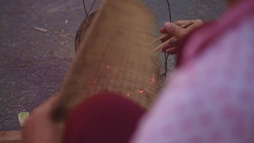 Woman cooks a flour tortilla over red hot coals outdoors using chopsticks and a fan