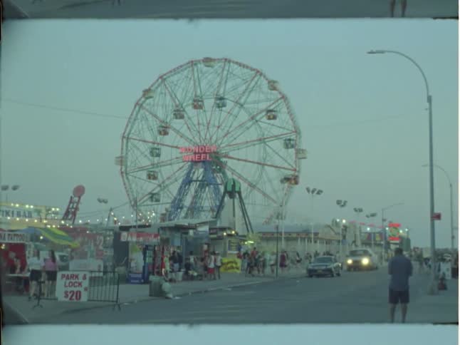 A ferris wheel looms of a carnival ground as people walking around it at sunset