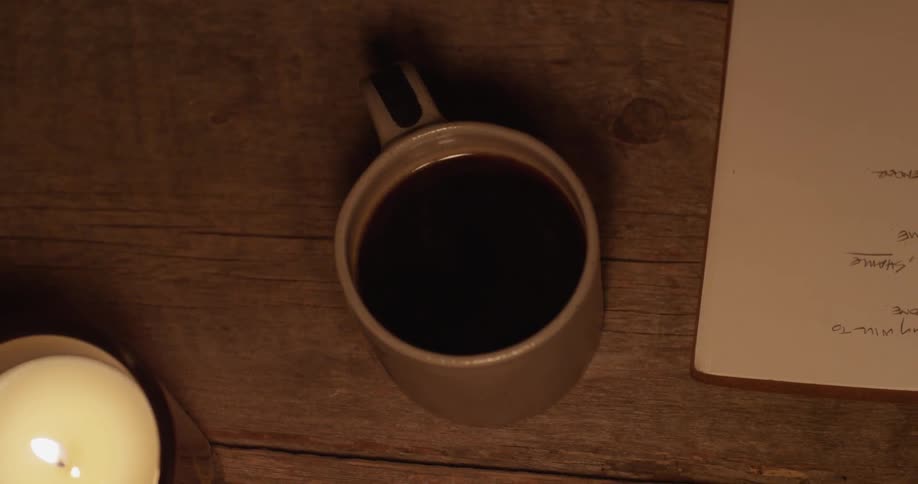 A man sets a cup of coffee on a desk while writing on paper