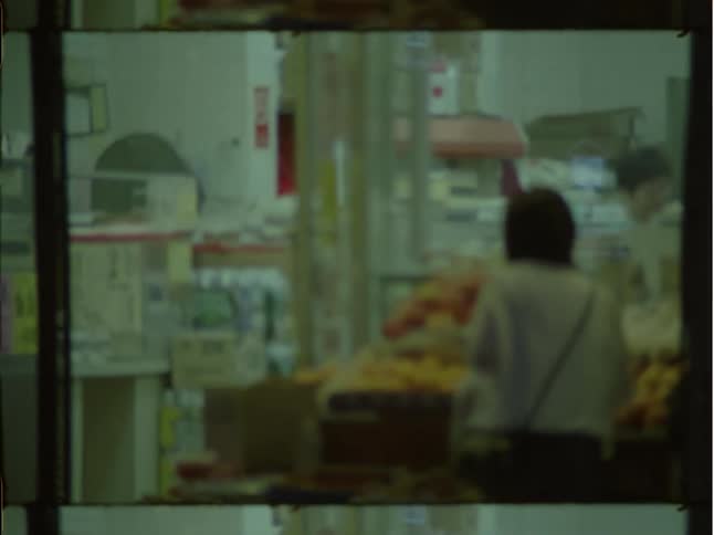 Customers examine produce in front of store as pedestrians walk by toward intersection corner