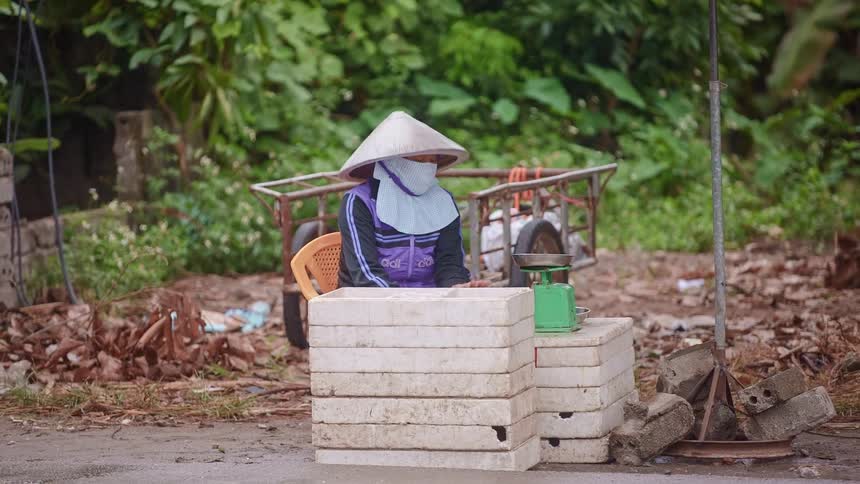 A person in a large hat and face covering sits by the side of a busy road behind a stack of bricks