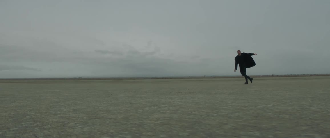 Man walking against what looks like a strong wind across a flat field on a cloudy day