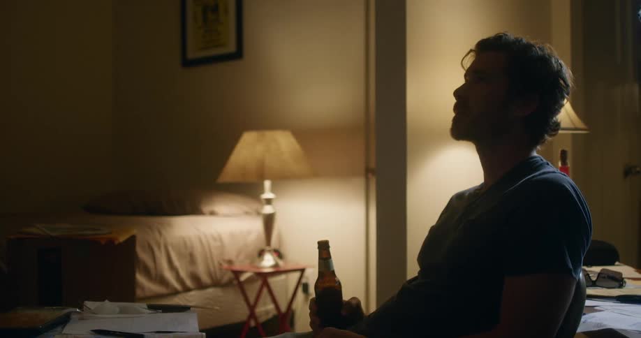 A man sitting on a chair holding a bottle with a bed and a lamp in the background